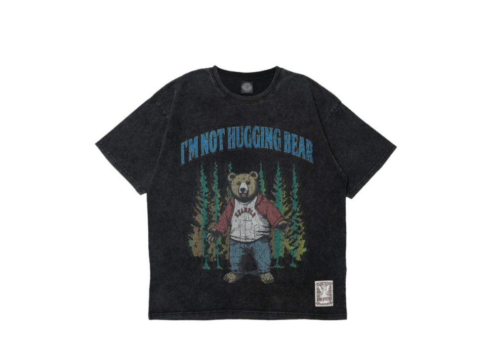 架空のストーリーをグラフィックに落とし込んだ、〈エグザンプル〉のTシャツ。こちらのTシャツはは、同ブランドで度々登場しているベア（熊）の、BB BEARをフィーチャー。森を彷徨うベアが、バーシティジャケットとデニムパンツを着用している。